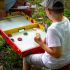 Kind spielt mit einem Erwachsenen ein Tischspiel mit Holzscheiben im Freien.